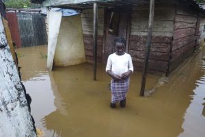 En flicka från Haiti som står utanför sitt hem i knä djupt brunt vatten efter stora mängder regn.